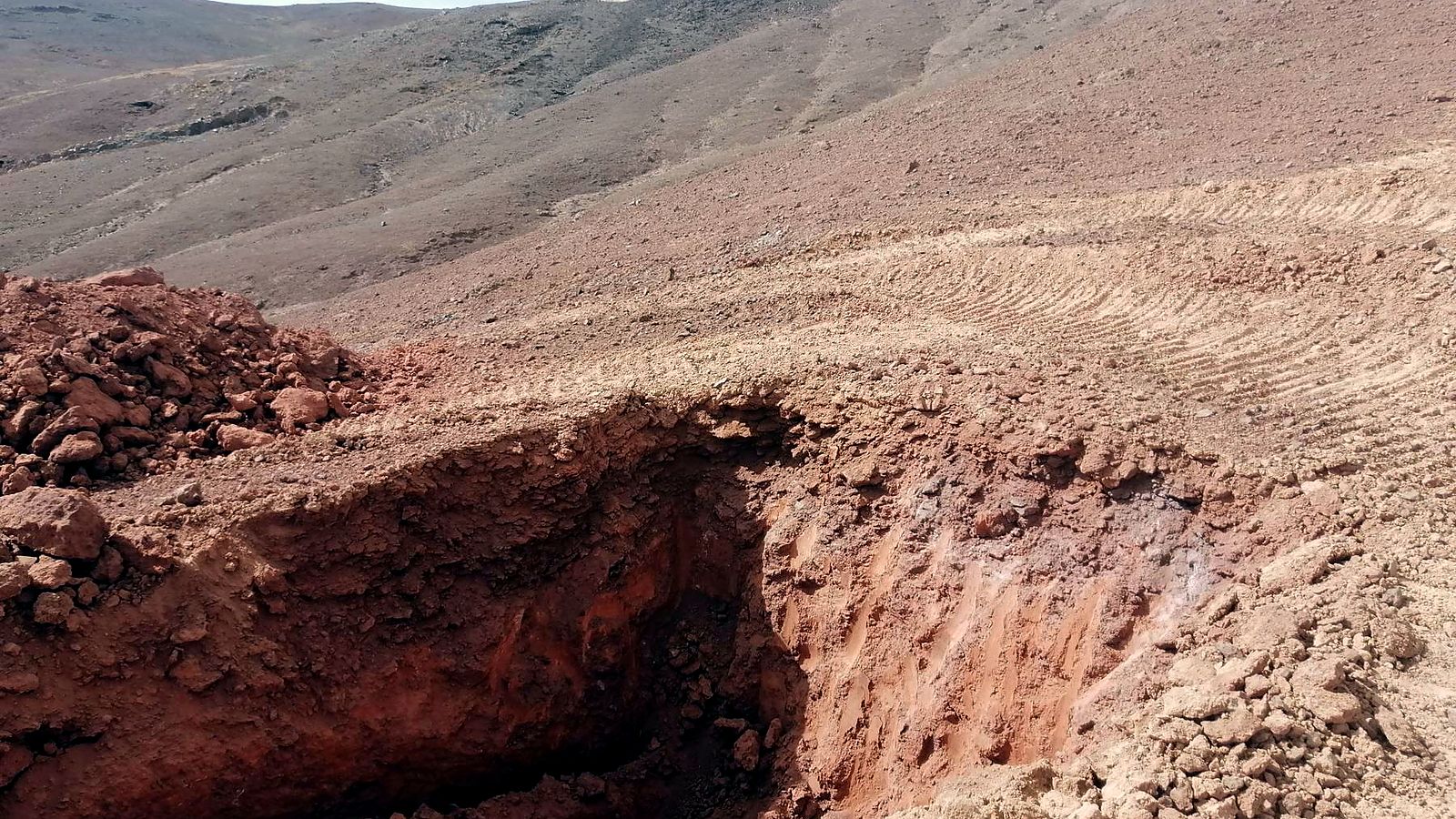 Exploration trench cut into reddish zeolitic tuff, with volcanic hills behind, at Ung Ar Rustom in northeastern Jordan